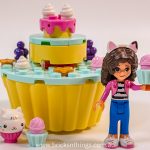 A LEGO-style figure with cat ears holds a pink cupcake, standing before a colourful, cupcake-shaped bakery from Gabby's Dollhouse. A small white cat and mini cupcakes add to the cakey fun on display.
