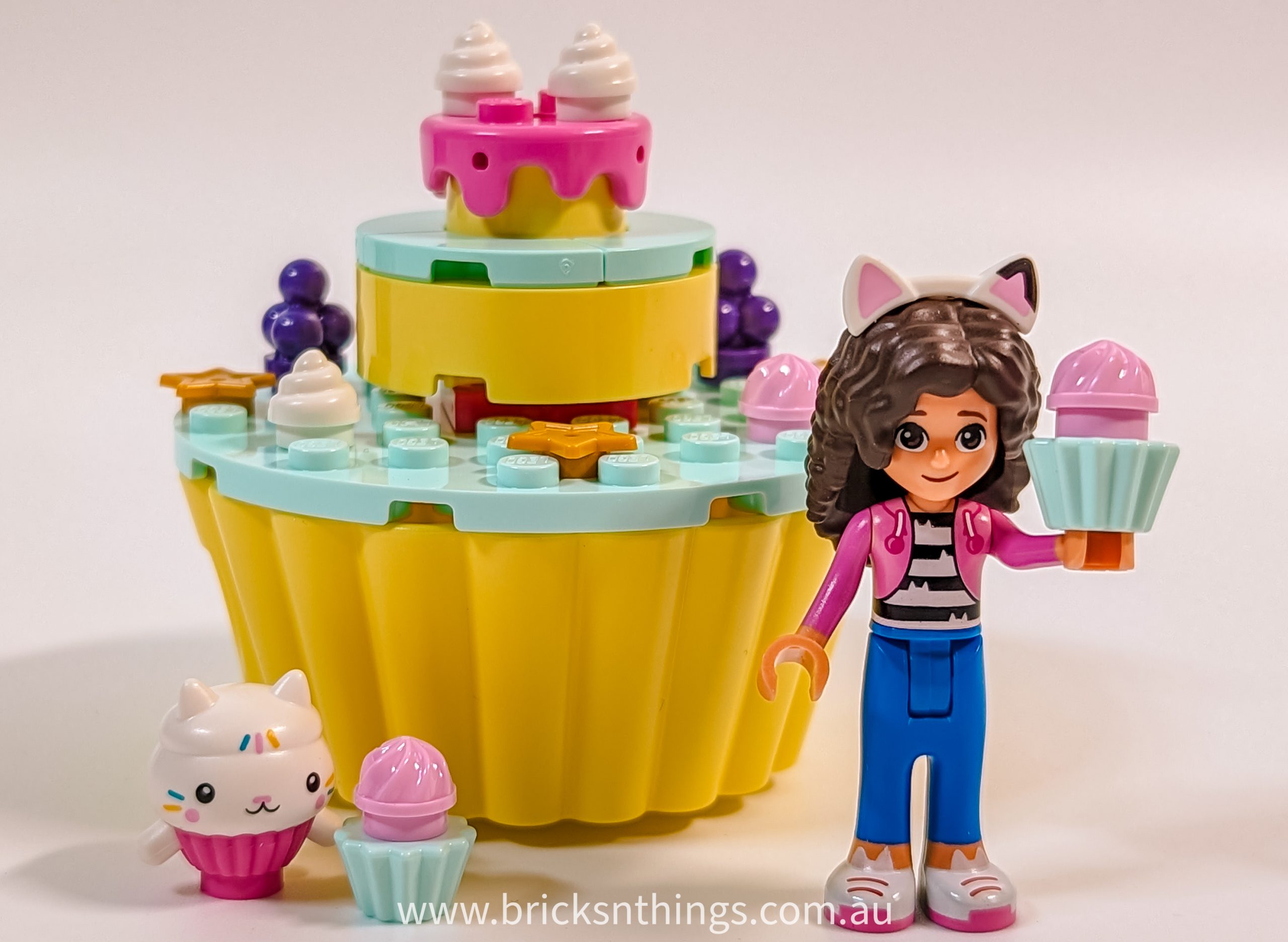 A LEGO-style figure with cat ears holds a pink cupcake, standing before a colourful, cupcake-shaped bakery from Gabby's Dollhouse. A small white cat and mini cupcakes add to the cakey fun on display.