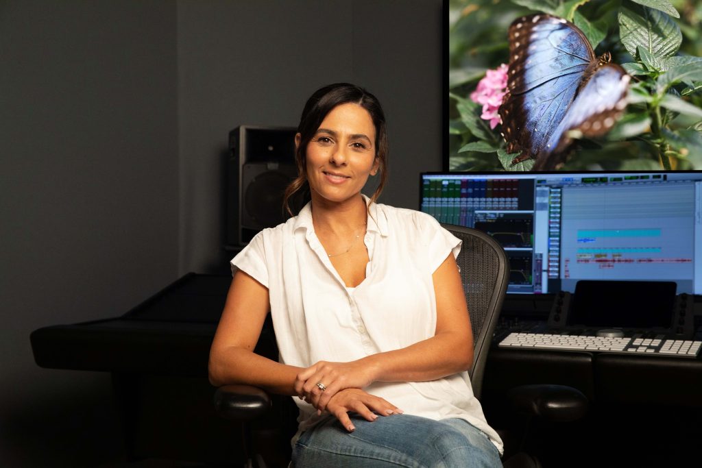 A woman in a white shirt sits in a recording studio, smiling at the camera. Behind her are computer screens, audio equipment, and a large image of a blue LEGO Insect butterfly on a green plant.