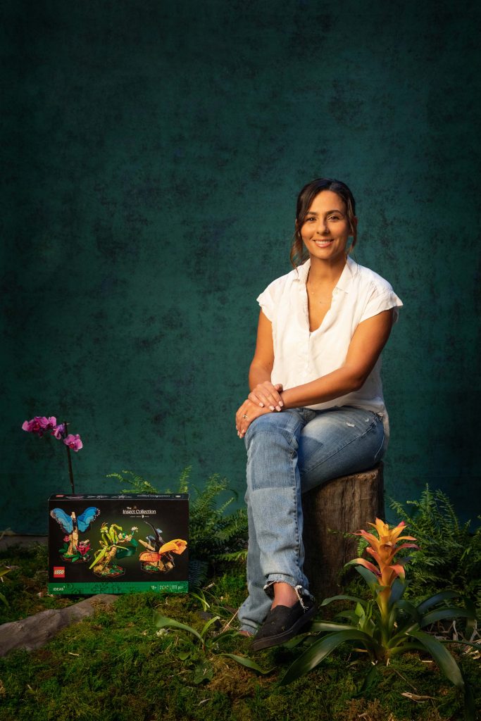 A woman in casual clothes sits on a tree stump, smiling, with a LEGO Insect box set and flowers arranged around her against a green, studio-style backcloth.