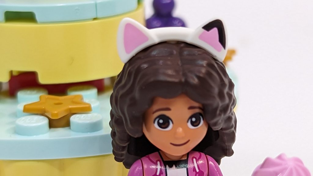 A LEGO minifigure with dark curly hair, wearing a white headband with pink cat ears, stands in front of a pastel-coloured LEGO bakery from Gabby's Dollhouse, complete with star-shaped decorations for extra Cakey Fun.