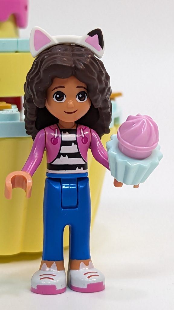 A LEGO-style mini-doll inspired by Gabby’s Dollhouse, featuring dark curly hair, a cat-ear headband, a pink jacket, blue trousers, and holding a cupcake with pink icing for extra Cakey Fun.