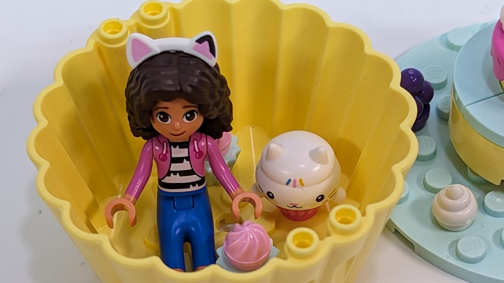 A LEGO-style mini-doll with curly hair, a pink jacket, and cat-ear headband sits in a yellow cupcake-shaped container beside a white cat figure and small cupcake toys.