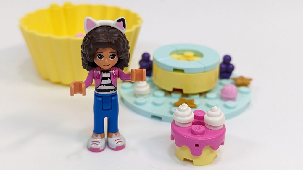 A LEGO mini-doll with curly hair, a pink jacket, cat ears—resembling Cakey from Gabby's Dollhouse—stands next to a pink cupcake and colourful toy desserts, with a yellow cupcake case in the bakery-themed scene.