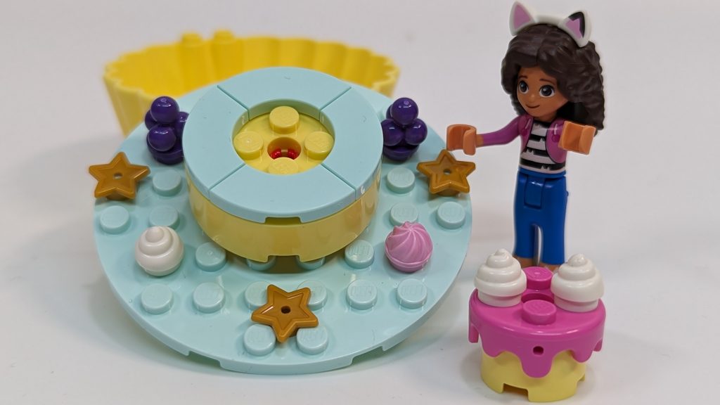 A LEGO-style mini-doll with cat ears stands next to pastel-coloured, cake-themed LEGO pieces decorated with star and swirl shapes, on a white background.