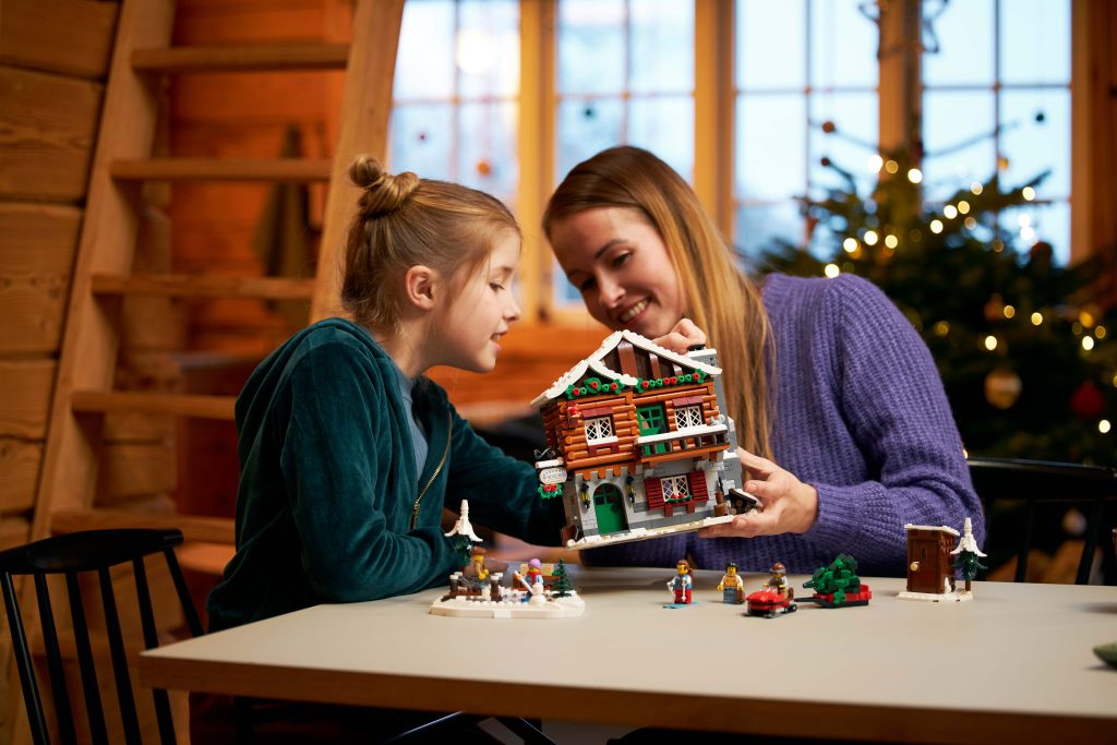 A woman and a girl sit at a table, smiling as they assemble the festive LEGO® Icons Alpine Lodge [10325] with toy figures scattered around. A Christmas tree glows in the background, adding magic to their winter holiday at home.