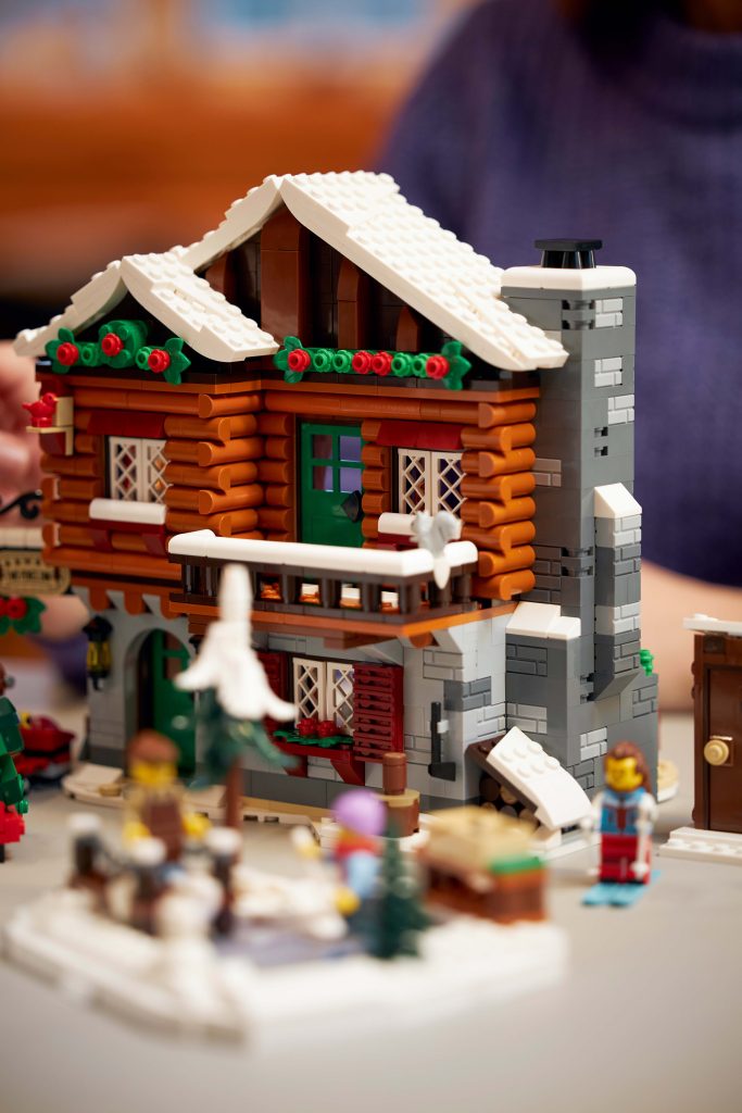 Take a Winter Trip to the LEGO® Icons Alpine Lodge [10325], a detailed winter cabin set with snow on the roof, festive greenery and red accents, surrounded by LEGO minifigures and charming snowy outdoor accessories.