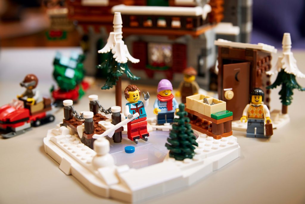 Take a Winter Trip to the LEGO® Icons Alpine Lodge [10325]: a charming winter scene with minifigures ice skating, pine trees, a wooden fence, and a snow-covered cabin in the background. One skater glides on the frozen pond whilst others watch in cosy winter clothes.