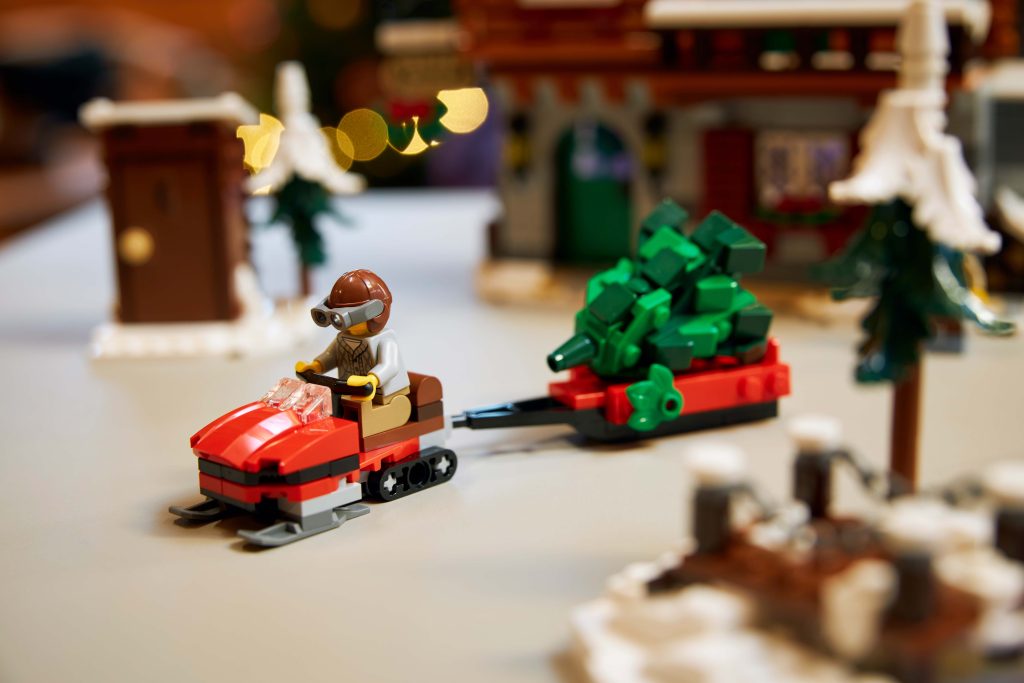 A LEGO figure takes a winter trip to the LEGO® Icons Alpine Lodge [10325], riding a red snowmobile and towing a green Christmas tree on a sledge through snowy buildings and festive winter scenery.