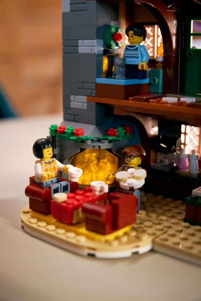 Two LEGO minifigures sit by a brick fireplace with mugs, surrounded by red sofas in the cosy LEGO® Icons Alpine Lodge [10325]. Another minifigure stands upstairs, completing this inviting winter holiday scene.