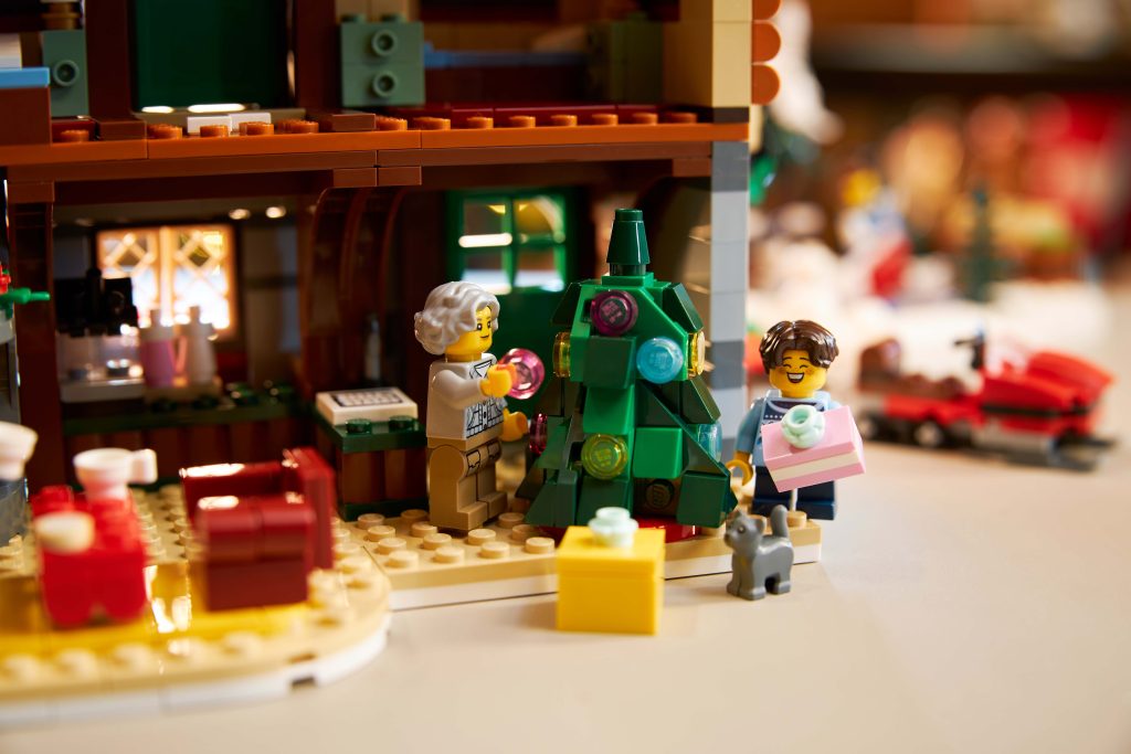 Two LEGO figures decorate a Christmas tree with colourful baubles in a cosy, festive scene at the LEGO® Icons Alpine Lodge [10325]; a grey cat and wrapped presents sit nearby inside the inviting LEGO house.