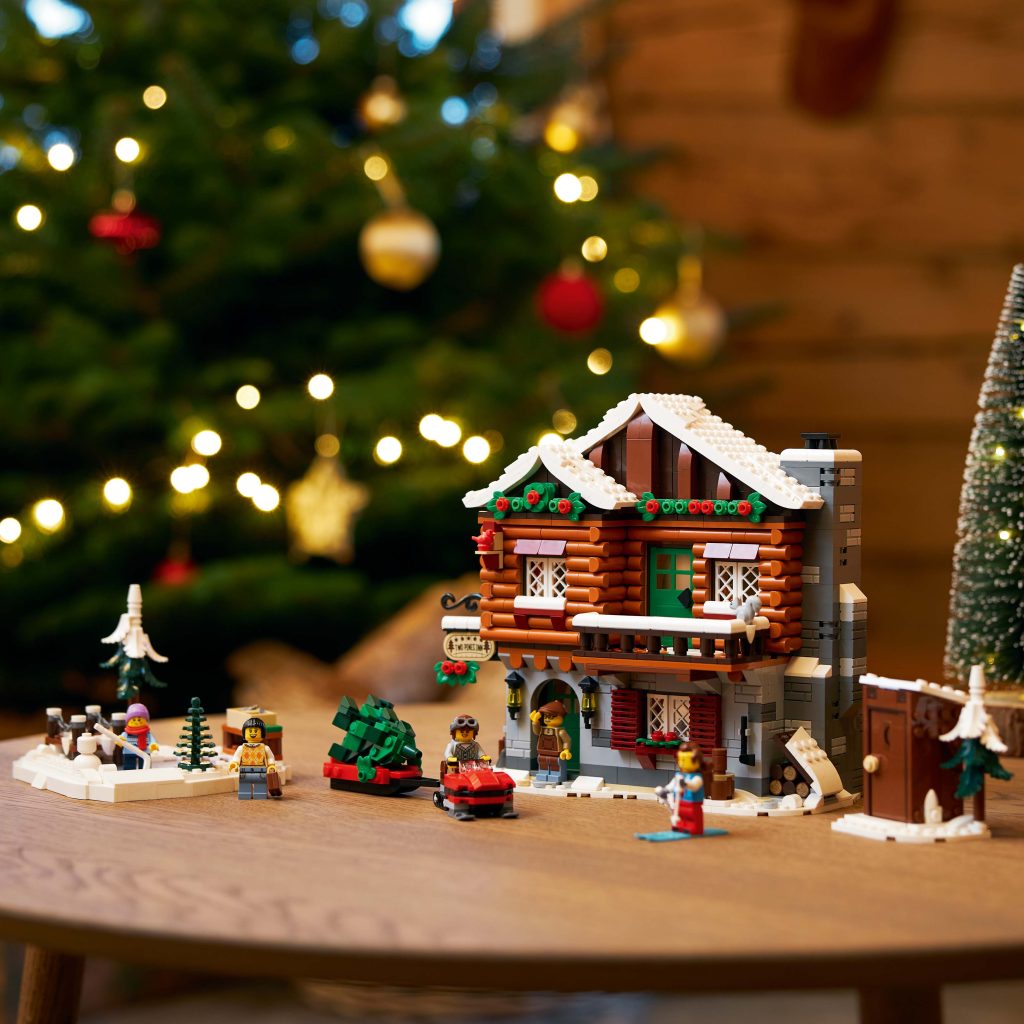 A festive LEGO log cabin, inspired by Take a Winter Trip to the LEGO® Icons Alpine Lodge [10325], sits on a table with minifigures, a car, trees, and holiday decorations. In the background, a glowing Christmas tree sparkles with yellow lights and baubles.