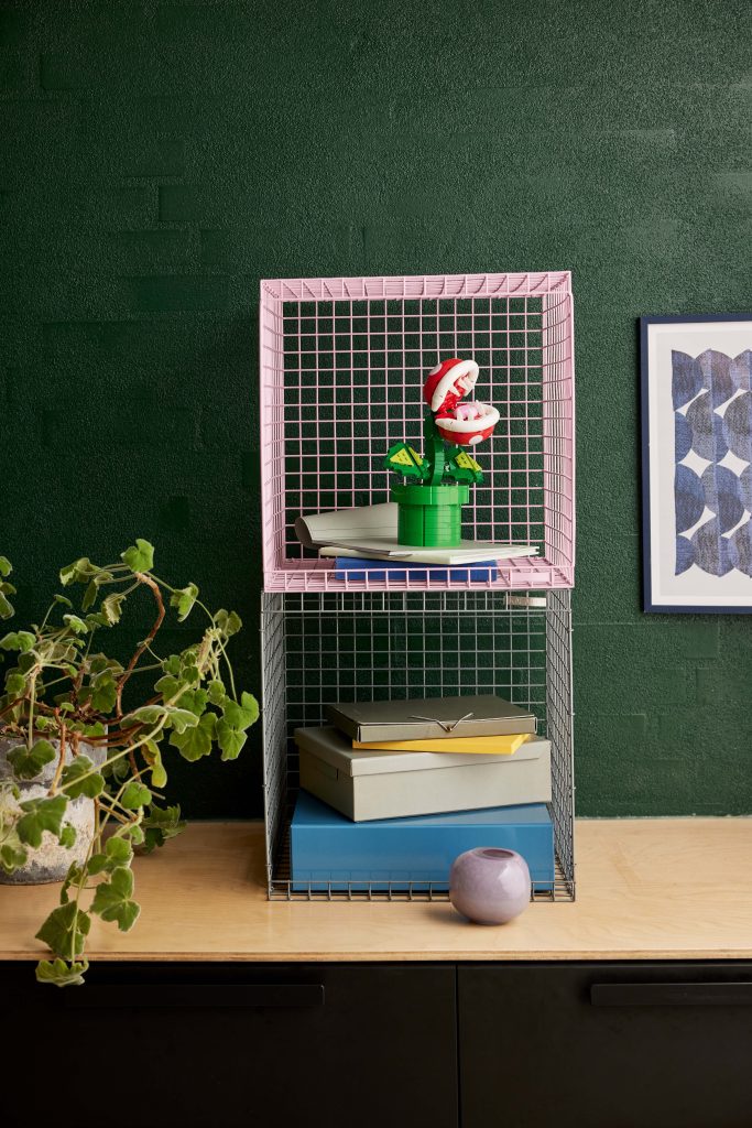 Two wire storage cubes, one pink and one silver, sit on a light wood surface against a dark green wall. Among boxes and books is a LEGO® Super Mario™ Piranha Plant [71426]. Nearby are a framed abstract artwork and another potted plant.
