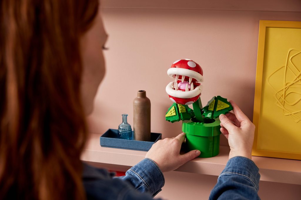 A person places the LEGO® Super Mario™ Piranha Plant [71426]—a red and white model—on a pink shelf, beside small decorative bottles and a yellow picture frame.