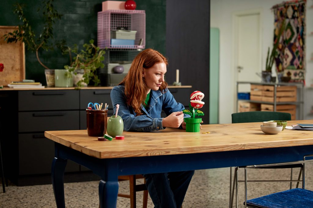 A woman with red hair sits at a wooden table in a colourful, modern kitchen, looking at the LEGO® Super Mario™ Piranha Plant [71426]. Art supplies and a bowl are on the table, with shelves, plants, and decor brightening the background.