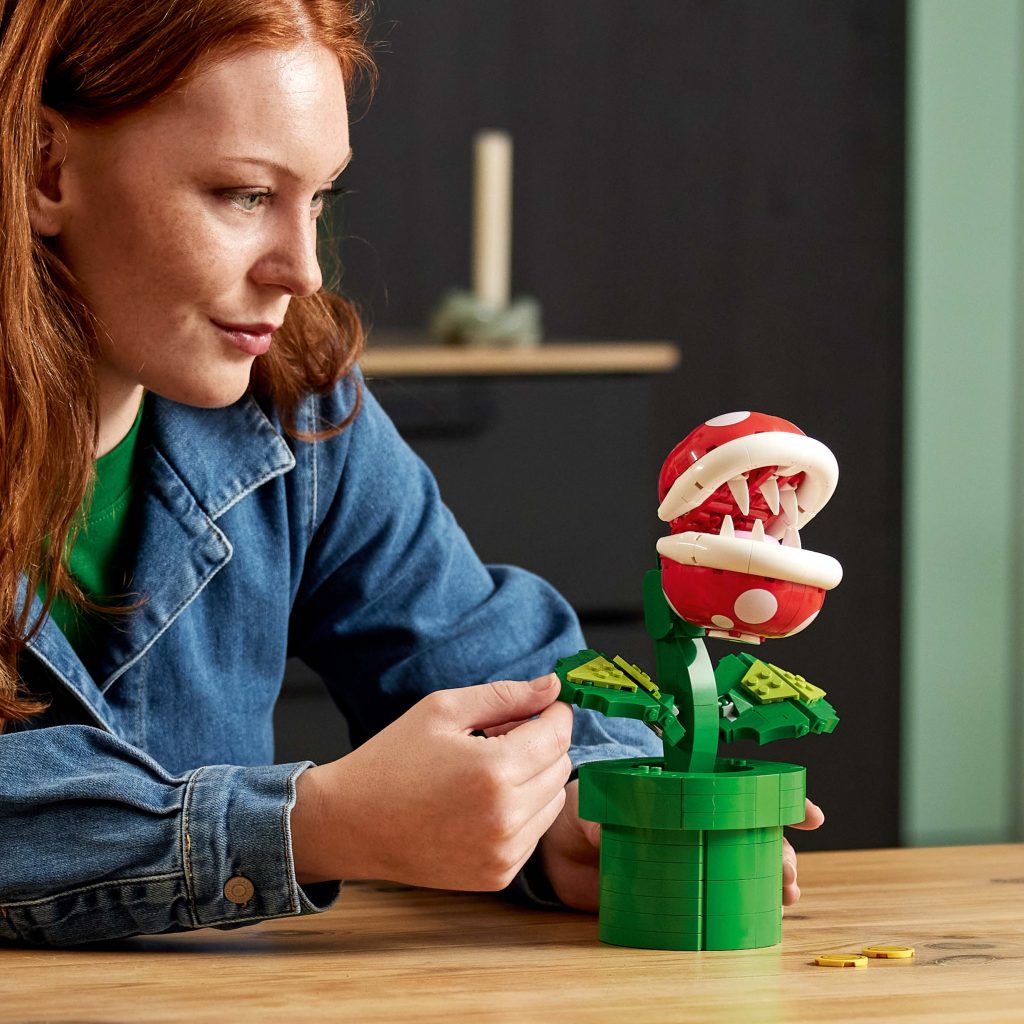 A person with red hair builds the LEGO® Super Mario™ Piranha Plant [71426], placing a green leaf on the red head with white spots and sharp teeth, as it sits in a green pot on a wooden table.