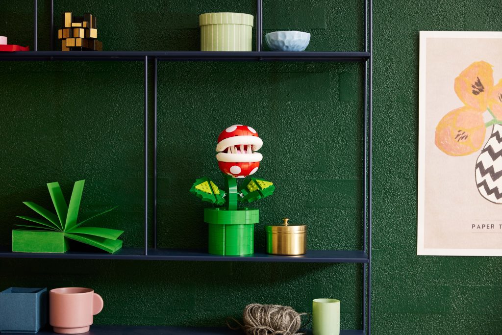 A LEGO® Super Mario™ Piranha Plant [71426] in red and white, with green leaves, sits on a dark shelf against a green textured wall, surrounded by stylish mugs, jars, and unique art pieces.