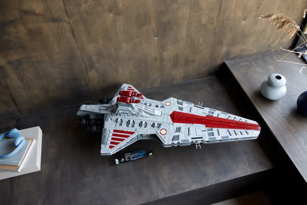 A large LEGO model of the Revealed: UCS Star Wars™ Venator-Class Republic Attack Cruiser with red and grey detailing is displayed on a dark surface beside a blue decorative object and two vases.