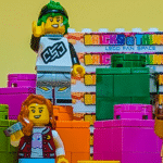 Colourful LEGO minifigures, including a witch, a bear, and others, stand among large LEGO Bricks n Things bricks in front of a yellow background and a stack of Bricks N Things signs.