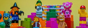 Colourful LEGO minifigures, including a witch, a bear, and others, stand among large LEGO Bricks n Things bricks in front of a yellow background and a stack of Bricks N Things signs.